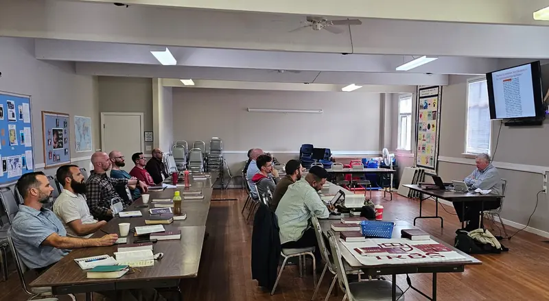 Students seated at long tables with open books during a class session, an IRBS banner on the table beside them